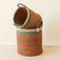 The LIKHA Abaca Cylinder Basket with handles, nested set of 2, features ethically made baskets in green and natural stripes, crafted from Manila hemp. One is tilted inside the other on a light wooden surface against a plain background.