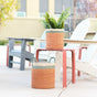 Two LIKHA Abaca Cylinder Baskets with handles, a nested set of 2 crafted from Manila hemp, feature green and blue accents; one holds a fern on a red table outdoors, while the other is placed near white and gray Adirondack chairs.
