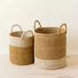 The LIKHA Abaca Cylinder Basket with handles, a nested set of 2, rests on a light wooden surface. Crafted from natural abaca fiber, these baskets feature tan hues with white stripes, adding artisan charm to any space.