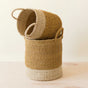 The LIKHA Abaca Cylinder Basket with handles, a nested set of 2, features natural and light beige Manila hemp and artisan charm, displayed with one basket upright and the other resting diagonally inside it on a light wooden surface.