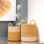 The LIKHA Abaca Cylinder Basket with handles, nested set of 2, features yellow and white color blocking. The smaller basket holds eucalyptus branches; both display artisan charm with abstract art hanging behind them.