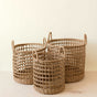The LIKHA Seagrass Rope Open Weave Cylinder Basket set features three handwoven baskets with sturdy handles, nested in varying sizes on a light wooden surface against a plain beige background.