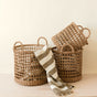 Three LIKHA Seagrass Rope Open Weave Cylinder Baskets with handles (nested set of 3) are shown on a light surface, two stacked and one upright. A striped beige and white fringed cloth drapes over the largest basket. Neutral background.