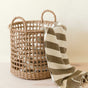 The LIKHA Seagrass Rope Open Weave Cylinder Basket with handles, from a nested set of 3, sits on a light wooden surface and holds a beige and white striped blanket draped over its edge.