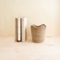 Three closed books with pale covers stand upright beside an Abaca Small Scallop Basket by LIKHA, ethically crafted from Manila hemp fiber, displayed on a light wood surface against a minimal, neutral background.
