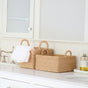 Two LIKHA Abaca Tapered Square Tabletop Baskets with Handles, handwoven by Filipino artisans, sit on a white bathroom counter holding folded towels. A pump bottle of liquid soap rests on a nearby tray.