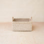 The LIKHA Abaca Square Tabletop Basket with Handles, handwoven from Abaca fiber and featuring two short handles, rests on a light wooden surface against a plain beige background.