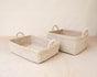 Two LIKHA Abaca Square Tabletop Baskets with handles, handwoven from natural straw and Abaca fiber, are displayed side by side on a light wooden surface against a neutral background.
