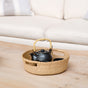 A black teapot with a yellow handle and cup sit on the LIKHA Abaca Round Tray with Cutout Handles, handcrafted from natural straw, atop a light wooden table with a beige couch in the background.