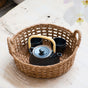 The Abaca Round Tray Open Weave with Cutout Handles from LIKHA holds a black teapot with a yellow handle and two matching black teacups, all set on a light wooden surface.