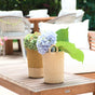 Two LIKHA Abaca Cylinder Vases, crafted from Manila hemp, display hydrangeas and greenery on a wooden outdoor table; cushioned chairs and a blurred patio are seen in the background.