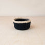The LIKHA Abaca Mini Trinket Basket, featuring a dark navy body and light beige rim, is handwoven using traditional techniques. It sits on a pale wooden surface against a neutral background.