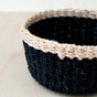 A close-up of the LIKHA Abaca Mini Trinket Basket, with a black body and light tan rim woven from abaca fiber, highlights traditional craftsmanship on a light wooden surface.