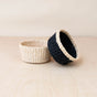 Two LIKHA Abaca Mini Trinket Baskets rest on a light wooden surface: one in natural beige, the other mostly black with a beige rim. The black basket leans against the beige, both set against a plain, neutral background.