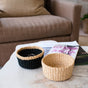 The LIKHA Abaca Mini Trinket Basket sits with another woven basket on a light wooden coffee table beside two magazines, with a beige sofa in the background—showcasing traditional craftsmanship and natural décor.