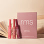 Three full-size RMS Beauty Serum Lipsticks from the Legends to Love Kit in pink and red stand upright by a matching pink box. An open book lies in the background on a neutral beige surface, showcasing this limited-edition lipstick set.