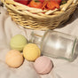 The Pacha Soap Co. Bath Bomb Set - Summer Garden features four pastel, natural ingredient bath bombs with an empty glass jar, displayed on light fabric with strawberries and an orange flower in the background.