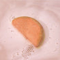 The Watermelon & Mint Bath Bomb by Pacha Soap Co. is a half-moon-shaped bath bomb that dissolves in pale pink, foamy water as nourishing plant oils and bubbles surround it.