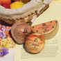 A Pacha Soap Co. Watermelon & Mint Bath Bomb, infused with nourishing plant oils, rests on an open book next to a wicker basket of lemons as sunlight casts gentle shadows on the scene.