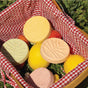 A basket lined with red and white checkered cloth holds Pacha Soap Co.'s Shower Steamers Set - Summer Garden, pastel-colored rounds inspired by garden scents, surrounded by lemons and apples on green grass.