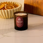 A lit Fall Signature Candle - Amber Glass by Pacha Soap Co. sits on a marble countertop with a bowl of dried orange slices in the background, filling the space with house-designed fragrances.
