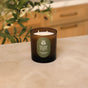 A brown amber glass Fall Signature Candle from Pacha Soap Co. rests on a beige countertop, with green leafy plants and wooden drawers softly blurred in the background—an inviting addition featuring house-designed fragrances for a uniquely curated scent experience.