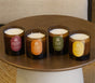 Four amber glass Fall Signature Candles from Pacha Soap Co., each with a differently colored label featuring a unique house-designed fragrance—Maplewood & Bergamot, Spiced Pumpkin & Amber, Fig Leaf & Pear, and Golden Spice & Chai—are arranged on a round wooden table. Crafted for a clean burn from a soy coconut apricot blend, these inviting candles infuse your space with distinctive autumnal aromas.