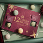 A maroon 12 Days Of Baths Advent Calendar with twelve perforated compartments for hand-crafted bath bombs, featuring gold text and star accents, surrounded by loose moisturizing bath bombs and ribbons.