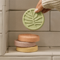 A hand holds a round, green, leaf-patterned Shower Steamer Set - Heritage Holiday Collection shower steamer above a stacked trio of orange, pink, and yellow aromatic shower steamers on a tiled ledge.