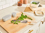 A kitchen scene features the Caraway Birch Wood Board holding a chef’s knife and fresh parsley, surrounded by stackable food containers with lids and a striped kitchen towel, all set on a white countertop.