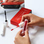A person holds Ere Perez Lash to Lip Party-Proof Duo—a red beetroot cheek & lip tint with white cap, beside its red box and black mascara wand—while a mirror in the background reflects the scene.
