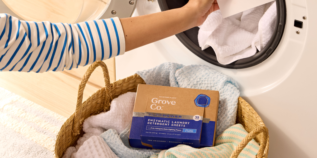 A person loads white towels into a front-loading washing machine. In the foreground, a wicker laundry basket holds folded towels and a box of Grove Co. Enzymatic Laundry Detergent Sheets.