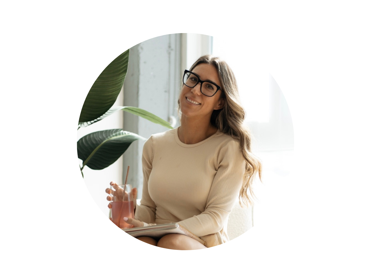 A woman with long wavy hair wearing glasses and a beige dress sits indoors, smiling and holding a glass of pink beverage with a straw, with a notebook on her lap and a large plant nearby.