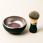 The Leaf Shave Shave Soap comes in a black and silver bowl for a creamy lather, paired with a shaving brush with a black handle and dense bristles, all displayed on a light surface.