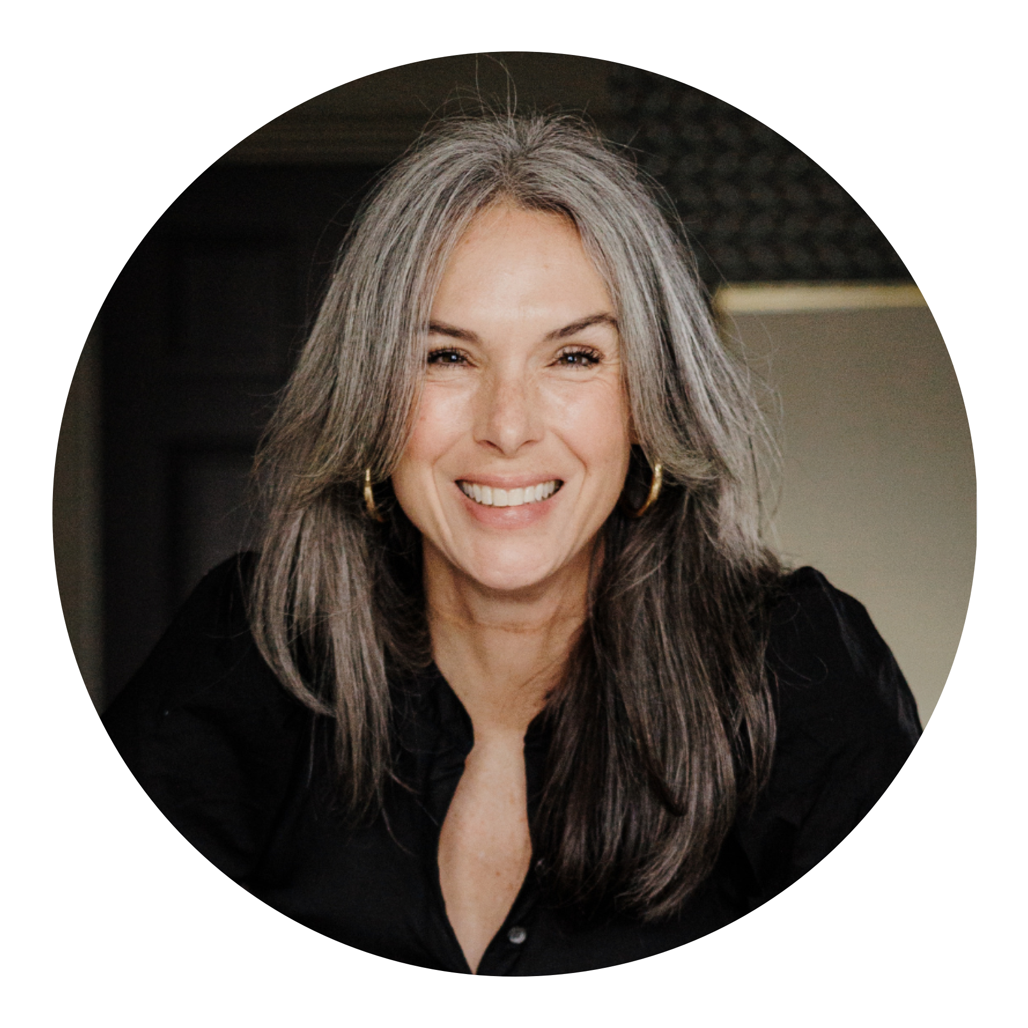 Smiling woman with long gray hair wearing a black top and gold hoop earrings, photographed indoors against a dark background.