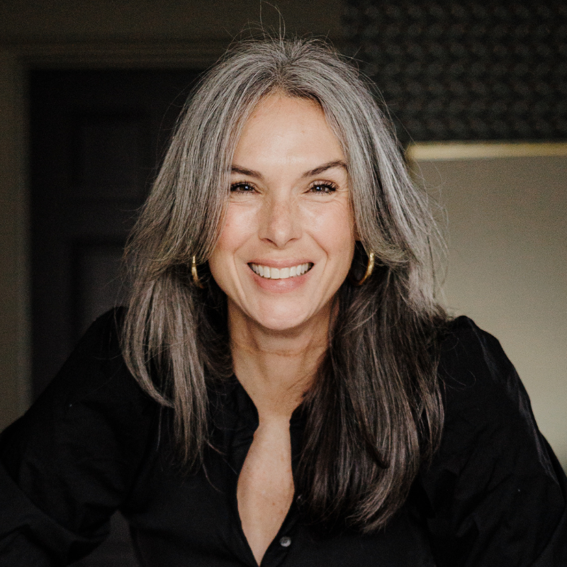 A woman with long gray hair, wearing a black shirt and gold hoop earrings, smiles warmly at the camera in a softly lit indoor setting.