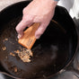 A hand uses the Lovett Sundries Cast Iron Scraper to remove stuck food residue from a black cast iron skillet.