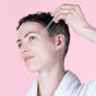 A person with short hair uses a dropper to apply Lovett Sundries Face Oil, enhancing skin elasticity. Theyre in a white robe with ear piercings against a soft pink background.