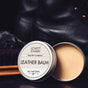 An open tin of Lovett Sundries Leather Balm sits next to a shoe brush and black leather boots. The light tan balm is labeled Seal & Condition, All Natural.