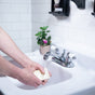 A person washes their hands with Lovett Sundries Pure Olive Oil Soap under running water in a white sink, while a small potted plant with pink flowers sits by the faucet on a tiled bathroom counter.
