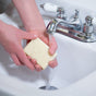 A person holds a Lovett Sundries Pure Olive Oil Soap bar under running water in a white sink, preparing to wash their hands—perfect for sensitive skin.