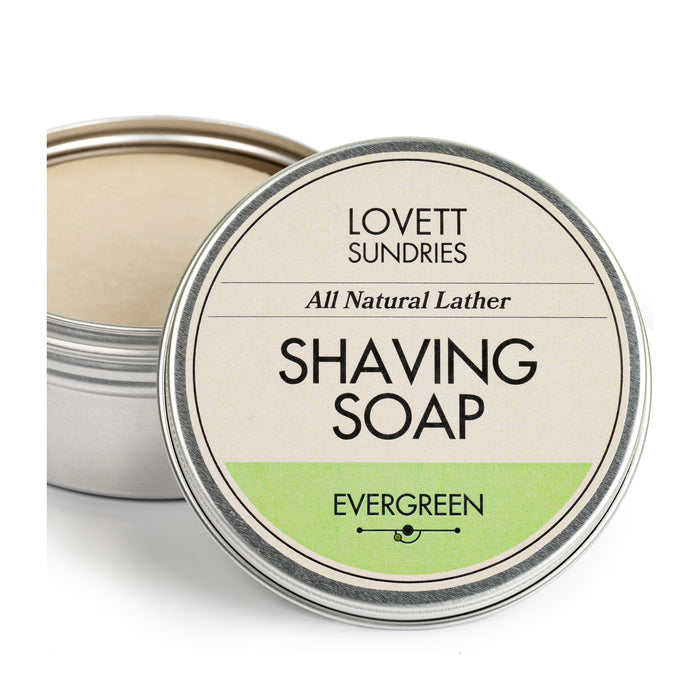 A tin of Lovett Sundries Shaving Soap showcases a lid with All Natural Lather and Evergreen, emphasizing its natural ingredients. The open tin reveals a smooth, light-colored soap enriched with bentonite clay.