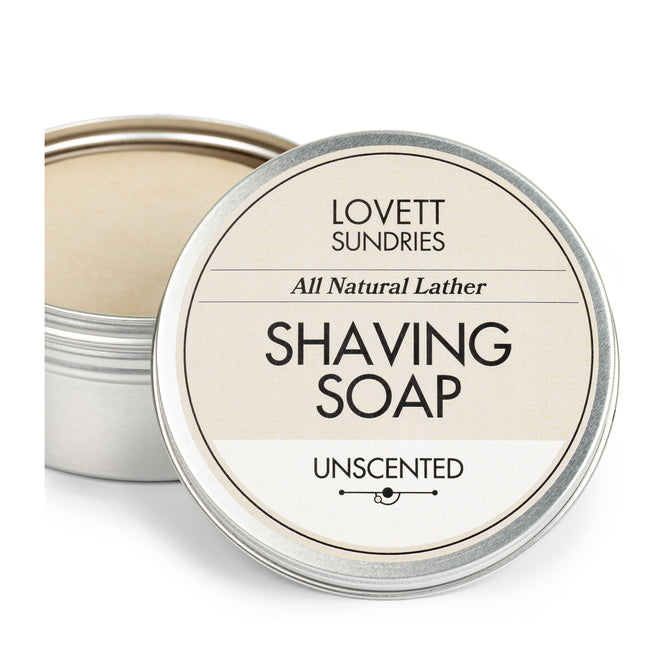 A round open tin of Lovett Sundries Shaving Soap is seen with its lid by its side, displaying the product name and brand in black text on a cream backdrop. Made from natural ingredients, it promises a smooth shave every use.
