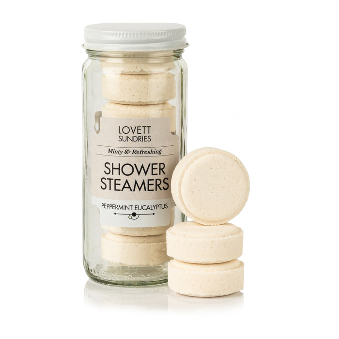 A jar of Lovett Sundries Shower Steamers contains round, beige tablets for aromatherapy with a soothing peppermint eucalyptus scent. Three extra tablets are stacked nearby.