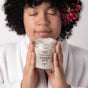 A relaxed person in a white robe, eyes closed and a flower in curly hair, holds a jar of Lovett Sundries Soaking Salts made with natural ingredients.