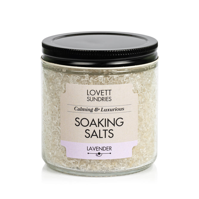A clear jar with a black lid contains Lovett Sundries Soaking Salts. The label reads “Calming & Luxurious Soaking Salts, Lavender.” Off-white crystals made with natural ingredients provide a soothing aromatherapy experience.