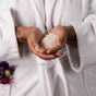 A person wearing a white bathrobe holds Lovett Sundries Soaking Salts in their cupped, tattooed hands, with purple and pink flowers in their pocket—reflecting the calming blend of natural ingredients.
