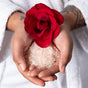 Tattooed hands in a white robe hold Lovett Sundries Soaking Salts with a red rose on top, evoking spa-like self-care with natural aromatherapy ingredients.