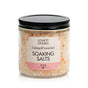 A clear jar with a black lid holds Lovett Sundries Soaking Salts. The label, accented in pink and beige, describes them as calming and luxurious. Inside are natural pink and white soaking salt crystals.