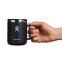 A hand holds a sleek black Hydro Flask Stainless Steel Mug, 12 oz, featuring a stainless steel rim and logo of a stylized person with raised arms. The mug, complete with a splash-resistant lid, is set against a pristine white background.